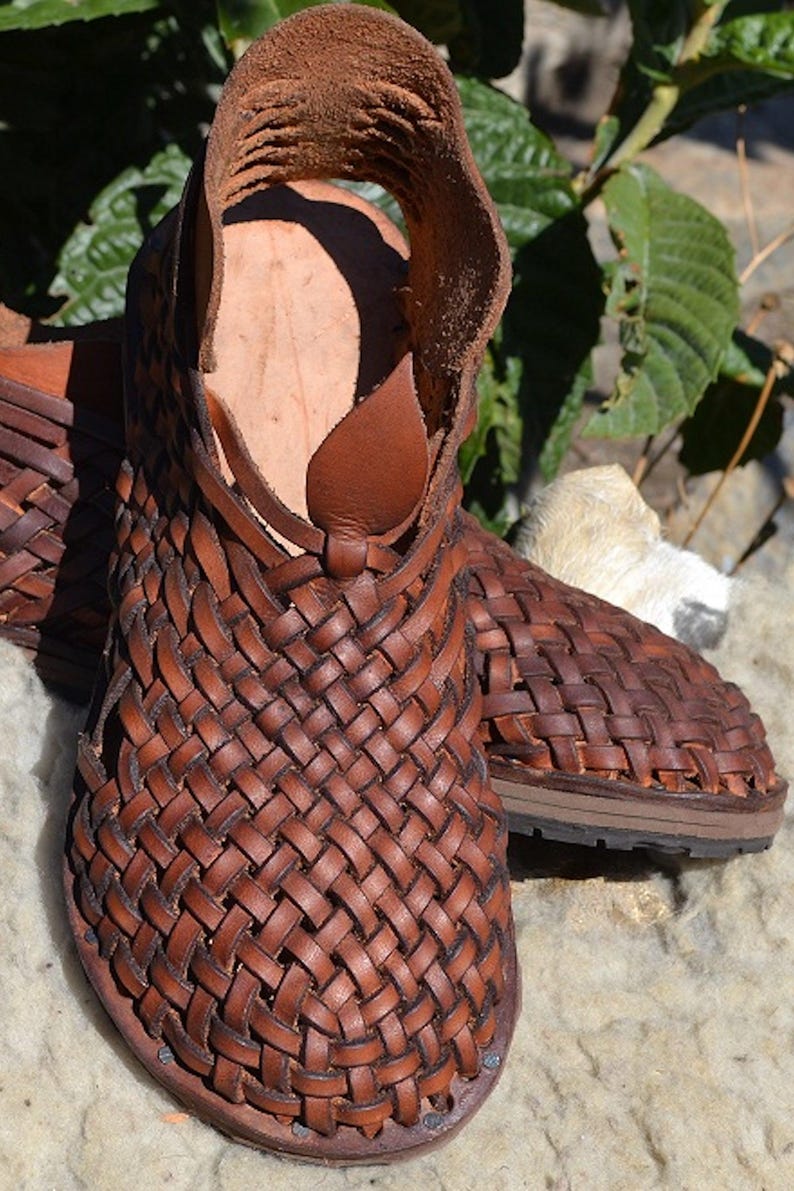 May include: A pair of brown leather sandals with a woven design. The sandals have a strap that goes around the ankle and a buckle closure.