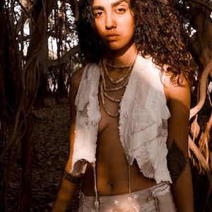 May include: A woman wearing a white, sleeveless, patchwork vest and skirt, adorned with shell accents. The outfit is paired with layered beaded necklaces. The background features a forest with large trees and hanging roots. The image has a warm, earthy tone.