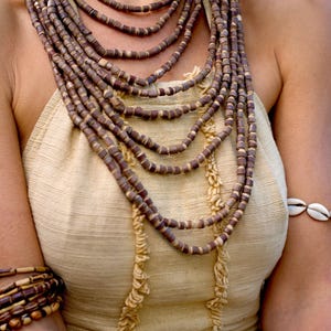 May include: A layered necklace made of brown wooden beads. The necklace is worn by a person with a light brown top and a shell bracelet on their wrist.