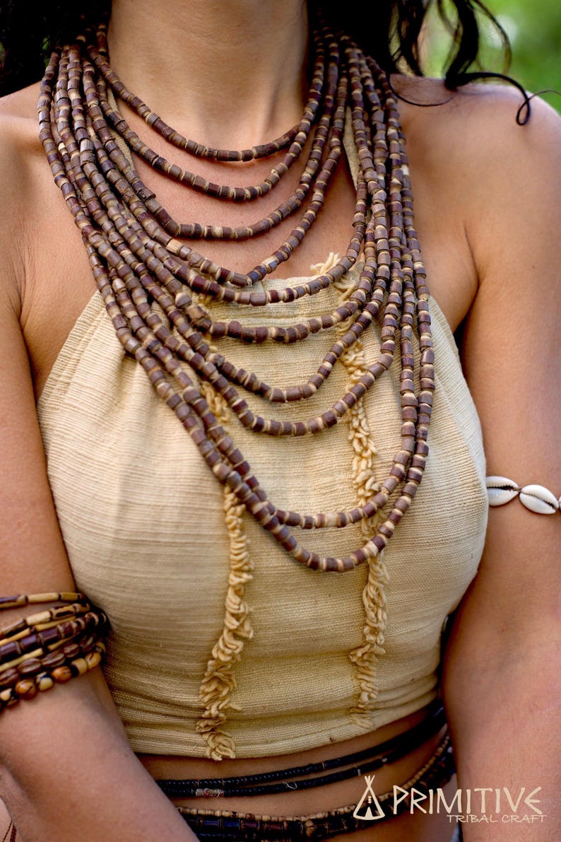 May include: A layered necklace made of brown wooden beads. The necklace is worn by a person with a light brown top and a shell bracelet on their wrist.