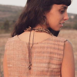May include: A woman wearing a sleeveless, light brown and white striped top with a brown cord necklace. The necklace has a small, brown, decorative bead at the end. The woman has long, dark brown hair and is wearing a hoop earring.