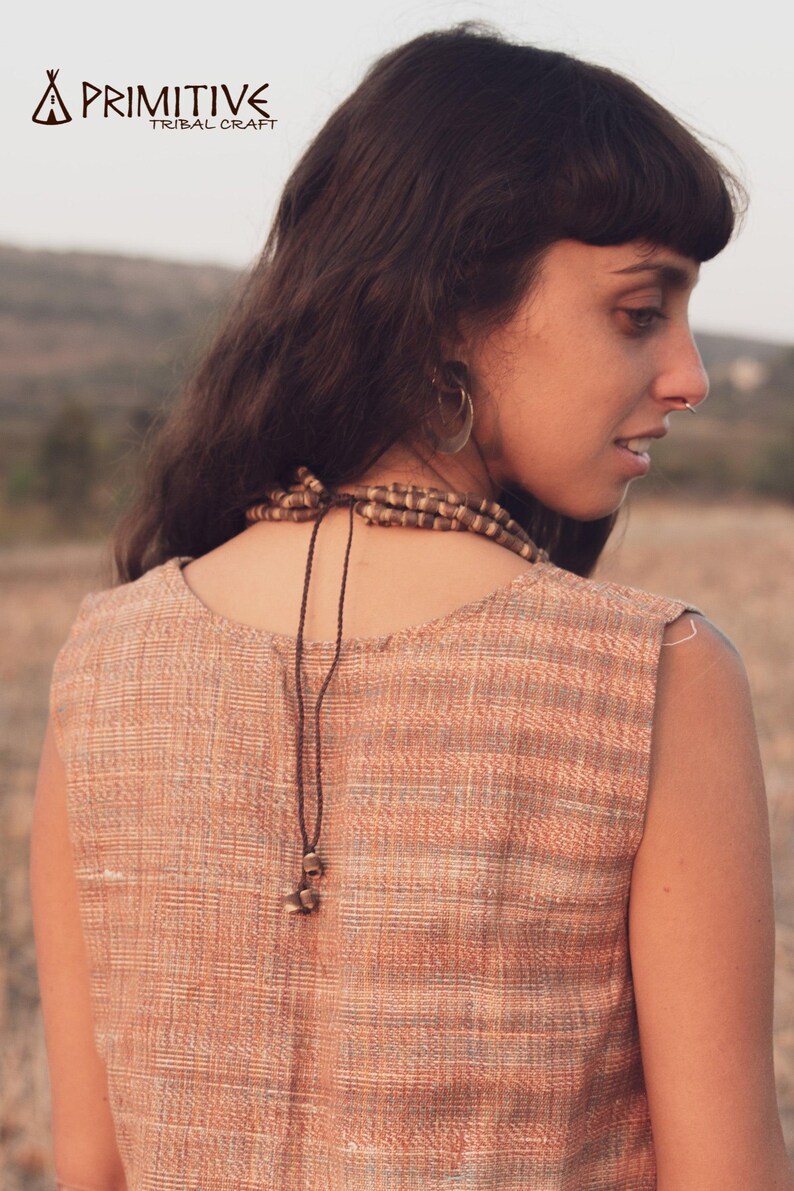 May include: A woman wearing a sleeveless, light brown and white striped top with a brown cord necklace. The necklace has a small, brown, decorative bead at the end. The woman has long, dark brown hair and is wearing a hoop earring.