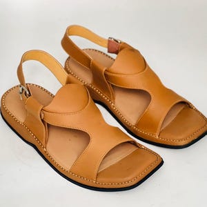 May include: A pair of tan leather sandals with a square toe design. The sandals have a strap that goes around the ankle and a strap over the top of the foot. The soles are black and the stitching is visible around the edges.