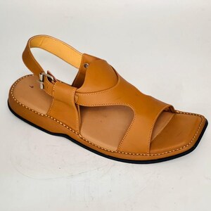 May include: A single, light brown leather sandal with a square toe and an adjustable ankle strap. The sandal features a stitched sole and a contrasting black outsole. The design includes a strap over the foot.