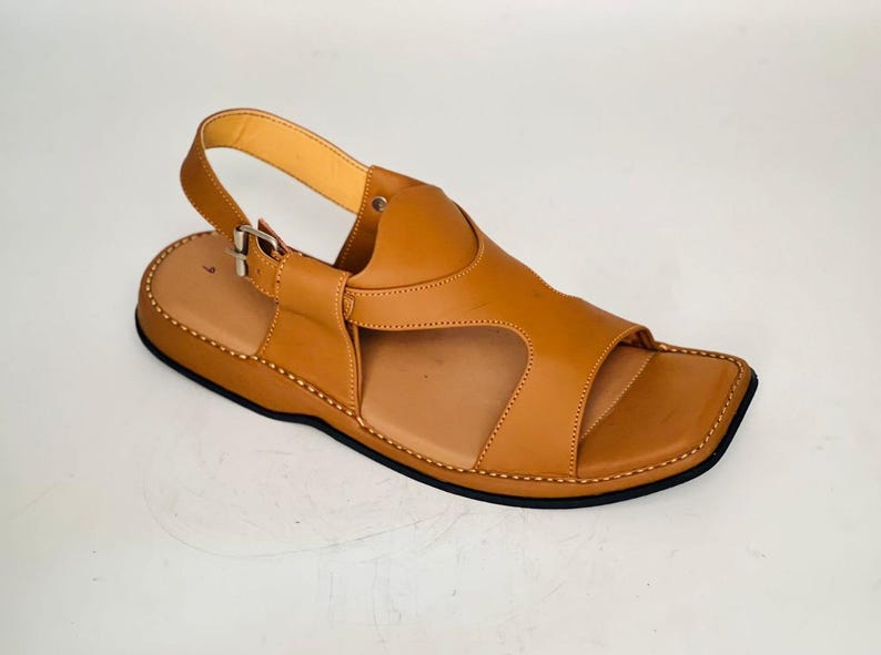 May include: A single, light brown leather sandal with a square toe and an adjustable ankle strap. The sandal features a stitched sole and a contrasting black outsole. The design includes a strap over the foot.