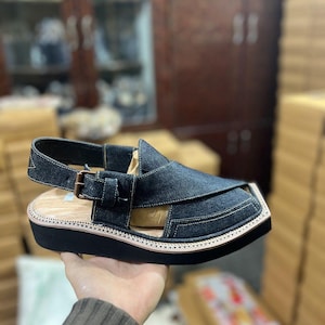 Pode incluir: Uma sandália de ganga azul-escura com design de biqueira fechada com painéis de tecido sobrepostos, tira lateral com fivela de metal cor de bronze, palmilha de madeira clara e sola grossa de borracha preta com costuras visíveis.