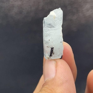 May include: A small, translucent, pale blue crystal specimen with a hexagonal prismatic shape. The crystal features a jagged, uneven top edge and contains a distinct, thin, dark black needle-like inclusion visible within its clear structure.