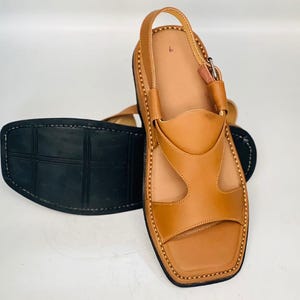 May include: Tan leather sandals with a square toe and a strap around the ankle. The sandals have a black sole and are stitched with a contrasting thread. The design includes cut-out details on the upper part of the sandal.