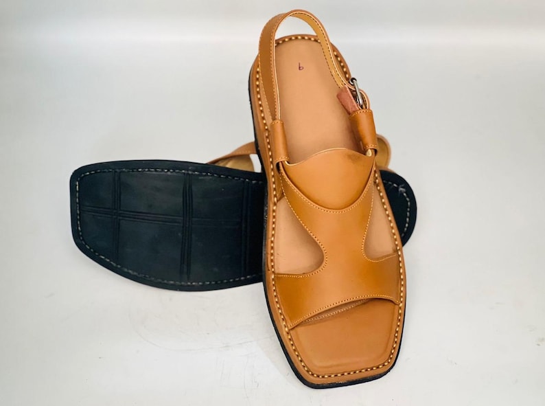 May include: Tan leather sandals with a square toe and a strap around the ankle. The sandals have a black sole and are stitched with a contrasting thread. The design includes cut-out details on the upper part of the sandal.