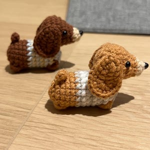 May include: Two handmade crochet dachshund dog figurines. One is brown and white, the other is tan and white. The dogs have black eyes and are positioned on a light wood surface.