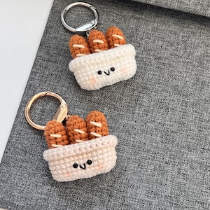 Pode incluir: Dois chaveiros de crochê com mini pães numa cesta. O pão é castanho com detalhes brancos, e a cesta é branco sujo com um rosto sorridente. Cada chaveiro tem um anel prateado ou dourado.