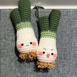 Peut inclure: Deux porte-clés en forme de poireaux crochetés à la main. Chaque porte-clés a un corps blanc, un dessus vert et un visage souriant avec des joues roses. Les porte-clés sont reliés par une chaîne en métal argenté.