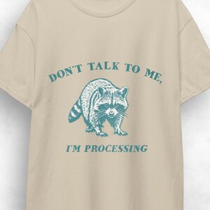 May include: Beige t-shirt with the text "DON'T TALK TO ME, I'M PROCESSING" in teal. A teal illustration of a raccoon is centered below the text. The t-shirt is hanging on a wooden hanger.