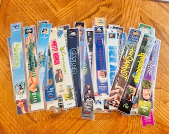 May include: A collection of VHS tapes, including titles like "Shrek," "Pretty Woman," and "Casper." The tapes are in their original plastic cases, with colorful labels and movie artwork visible. The tapes are arranged on a wooden surface.
