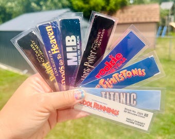 May include: A hand holding a collection of movie-themed bookmarks. The bookmarks feature titles like "Harry Potter," "Titanic," and "Cool Runnings." Each bookmark is in a clear plastic sleeve, with vibrant colors and various fonts.