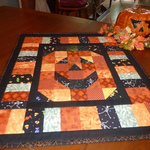 May include: A Halloween-themed table runner featuring a patchwork design with an orange jack-o'-lantern face at the center. The runner is bordered in black and includes various Halloween-themed fabric patterns in orange, black, and gray.