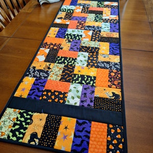 May include: A black, orange, purple, and green patchwork table runner with a Halloween theme. The runner features bats, pumpkins, candy corn, and ghosts.