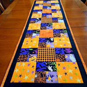 May include: A rectangular quilted table runner featuring a patchwork design of orange, purple, and black fabric squares. The pattern includes motifs of jack-o'-lanterns, ghosts, bats, spiderwebs, stars, and candy corn, finished with a black border.
