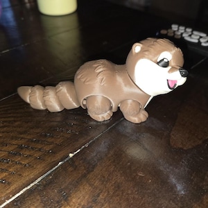 May include: A brown and white articulated otter figurine with a segmented tail. The otter has a smooth brown body, a white face, a small black nose, and a pink mouth. The figure features visible joints along the tail and body, allowing for movement and posing of the plastic animal toy.