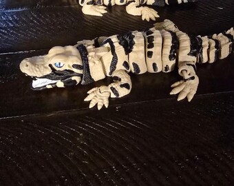 May include: Two toy alligators are displayed. They are tan with black stripes and accents. The alligators have blue eyes and open mouths. The toys are on a dark wooden surface.