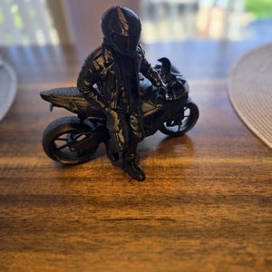 May include: A black 3D printed figurine of a person wearing a helmet and a jacket, leaning against a black sport motorcycle. The figure is standing beside the bike with one hand resting on the seat, showcasing detailed textures on the gear and wheels.