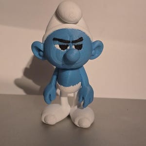 May include: A blue figurine of a Smurf character with a grumpy expression. The figure has a white hat and white shoes. The Smurf is standing on a gray surface.