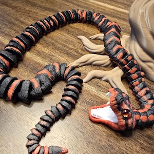May include: A segmented, articulated snake toy in black and orange. The snake's head is open, revealing a white interior and red mouth. The snake has a detailed, textured appearance, with a realistic design.