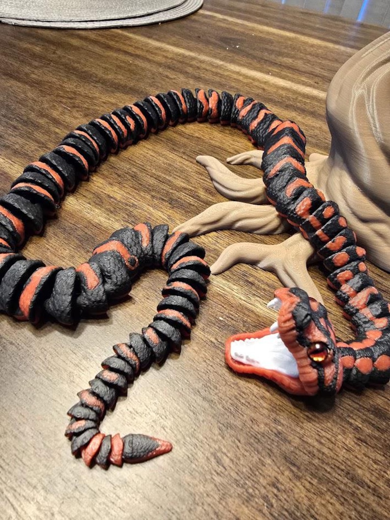 May include: A segmented, articulated snake toy in black and orange. The snake's head is open, revealing a white interior and red mouth. The snake has a detailed, textured appearance, with a realistic design.
