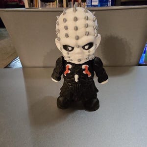 May include: A small, stylized figurine of a character with a white head covered in silver spikes. The figure has a black outfit with red accents and black boots. The figurine is a collectible item.