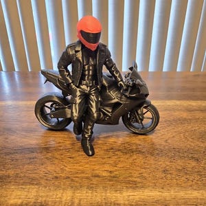 May include: A 3D printed figurine of a person wearing a black leather-style jacket and pants, leaning against a black sport motorcycle. The figure wears a bright red helmet with a black visor. The entire set is rendered in a monochromatic black finish.