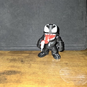 Puede incluir: Una figura negra de Venom con cabeza y dientes blancos y lengua roja. La figura est&aacute; sobre una superficie de madera.
