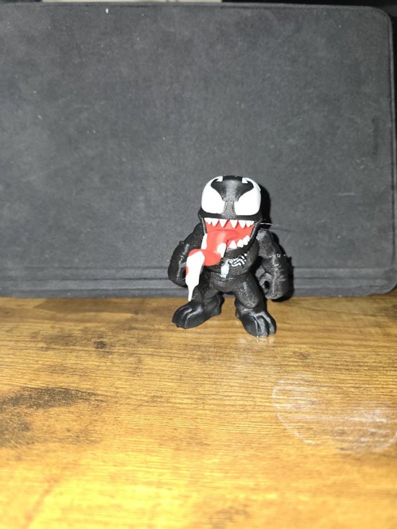 Puede incluir: Una figura negra de Venom con cabeza y dientes blancos y lengua roja. La figura est&aacute; sobre una superficie de madera.