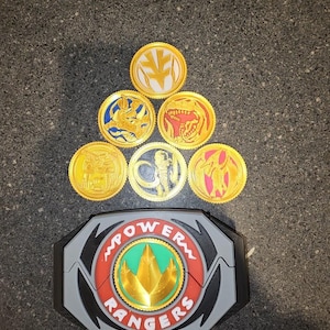 May include: A gray Power Rangers belt buckle with a central green and gold emblem, accompanied by six circular gold-toned coins featuring various embossed animal designs including a white tiger, triceratops, tyrannosaurus, mastodon, and pterodactyl.