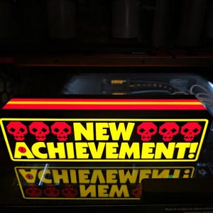 May include: A rectangular light box with a black frame and a glowing yellow front panel. The text NEW ACHIEVEMENT! is printed in bold yellow letters, flanked by three red pixelated skull icons on each side. A red horizontal stripe runs across the top.