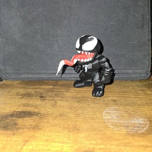 Puede incluir: Una figura de Venom en blanco y negro con lengua roja y dientes blancos. La figura est&aacute; sobre una superficie de madera. La figura tiene un emblema de ara&ntilde;a blanco en el pecho.