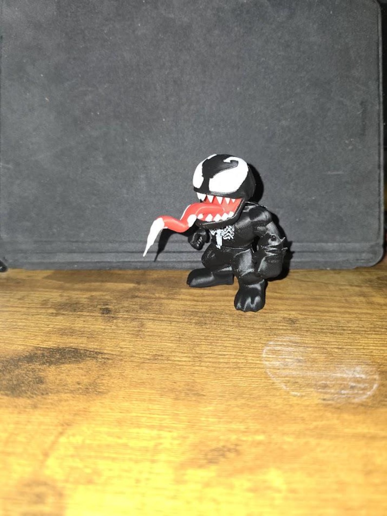 Puede incluir: Una figura de Venom en blanco y negro con lengua roja y dientes blancos. La figura est&aacute; sobre una superficie de madera. La figura tiene un emblema de ara&ntilde;a blanco en el pecho.