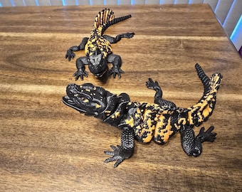 May include: Two toy lizards with black bodies and yellow and orange patterns. The lizards are posed on a wooden surface. One lizard is larger and in the foreground, while the other is smaller and in the background.