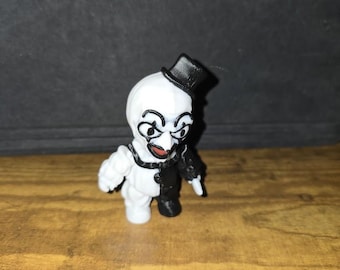 May include: A small, white and black figurine of a clown with a top hat. The clown has a painted face with exaggerated features, including large eyes, a red mouth, and a black and white outfit. The figurine is standing on a wooden surface.