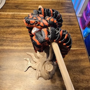 May include: A 3D-printed sculpture of a snake with a red and black pattern. The snake is coiled around a wooden stick, and the base is a beige tree-like structure. The snake has orange eyes and is displayed on a wooden table.
