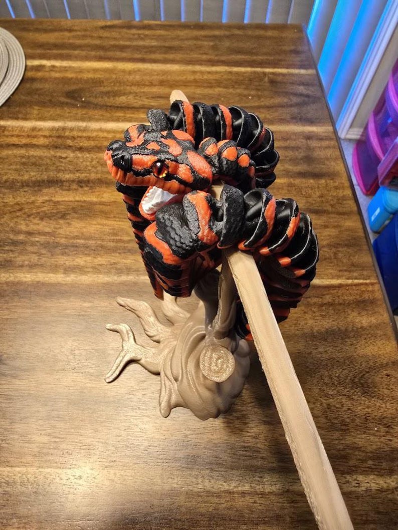 May include: A 3D-printed sculpture of a snake with a red and black pattern. The snake is coiled around a wooden stick, and the base is a beige tree-like structure. The snake has orange eyes and is displayed on a wooden table.