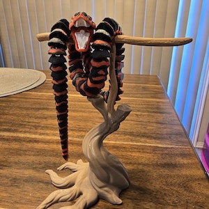 May include: A 3D-printed articulated snake sculpture in black and red stripes, posed on a light brown tree-like stand. The snake's mouth is open, revealing white teeth. The sculpture is displayed on a wooden table.