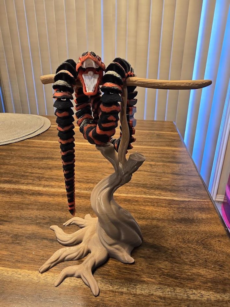 May include: A 3D-printed articulated snake sculpture in black and red stripes, posed on a light brown tree-like stand. The snake's mouth is open, revealing white teeth. The sculpture is displayed on a wooden table.