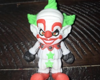 May include: A small, detailed clown figurine with a white head and body, green hair and accents, and a red nose and mouth. The clown has black gloves and red shoes. The figurine has a stern expression and is decorated with green star shapes.