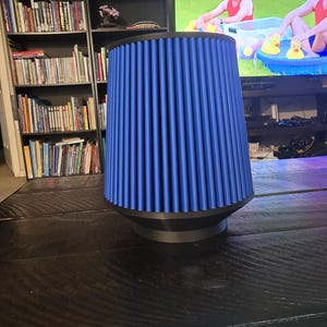 May include: A blue air filter with a black base. The filter has vertical ridges and a cylindrical shape. It is sitting on a dark wooden table. The background includes bookshelves and a television.