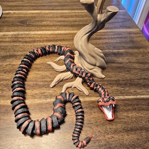 May include: A detailed sculpture of a snake with a red and black pattern, coiled and posed near a tree-like structure. The snake's mouth is open, revealing a white interior. The sculpture is set on a wooden surface.