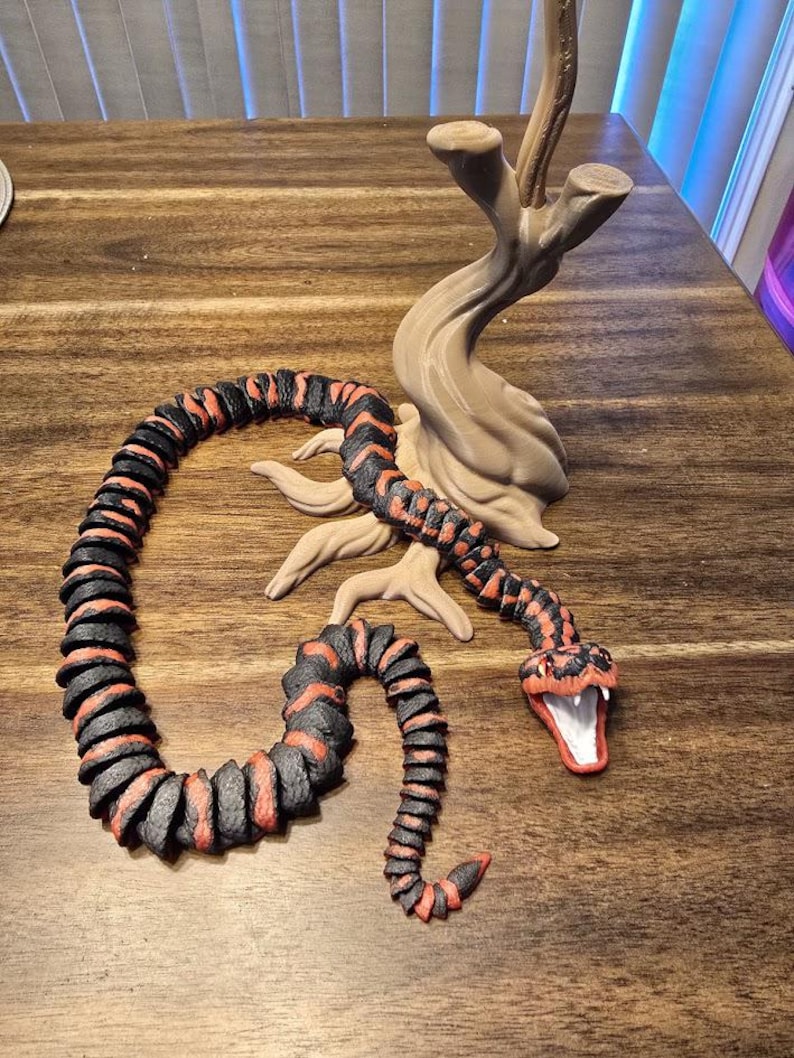 May include: A detailed sculpture of a snake with a red and black pattern, coiled and posed near a tree-like structure. The snake's mouth is open, revealing a white interior. The sculpture is set on a wooden surface.
