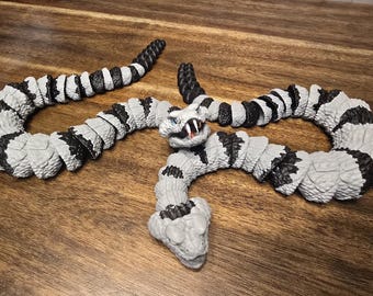 May include: A gray and black toy snake with a detailed design. The snake has a segmented body with black and white patterns. The head is gray with white fangs and blue eyes. The snake is posed on a wooden surface.