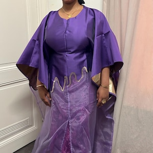 Peut inclure: Une robe violette et une coiffe assortie. La robe présente un corsage avec une bordure dorée et des manches fluides. La jupe a un voile transparent et un panneau en dentelle. La personne porte des bijoux en or.