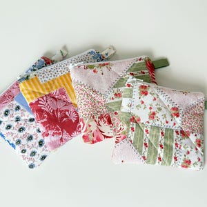 Peut inclure: Quatre pochettes en tissu matelassé avec différents motifs floraux et couleurs, dont le rouge, le rose, le bleu et le jaune. Chaque pochette a une fermeture éclair et une petite boucle en tissu. Disposition sur une surface blanche.