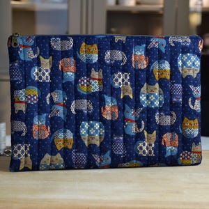 May include: A rectangular quilted zipper pouch in deep navy blue fabric featuring a repeating pattern of stylized cats in various colors and geometric prints. The pouch has vertical stitching lines and a metallic zipper pull on the left side.